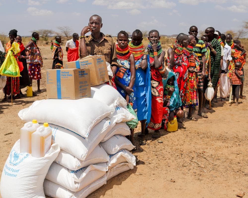 People queue behind large bags and boxes piled up outdoors